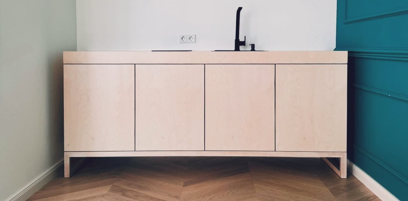 Helles Sideboard mit vier Türen auf einem schlanken Gestell vor einer Wand in Petrol